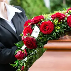 Osoba w czerni kładzie wieniec z czerwonych róż na trumnie. Czy wieniec jest konieczny? To symbol pożegnania.