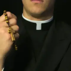 Dłoń księdza trzyma drewniany różaniec. Czy ksiądz może odmówić pochówku? Pytanie o sakramenty.