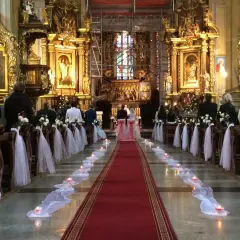Wnętrze kościoła udekorowane na ślub. Wzdłuż czerwonego dywanu stoją ławki ozdobione białymi kwiatami i tiulem. Widać świece i gości.