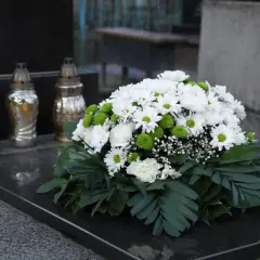 Białe i zielone kwiaty tworzą nowoczesne dekoracje na pomnik. Obok stoją trzy znicze.