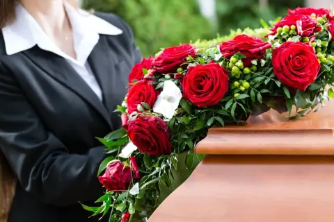 Osoba w czerni kładzie wieniec z czerwonych róż na trumnie. Czy wieniec jest konieczny? To symbol pożegnania.
