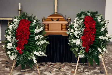 Dwa wieńce pogrzebowe z czerwonych róż i białych kwiatów stoją obok drewnianej trumny. Czy na kremację kupuje się kwiaty? Tak, to piękny gest.