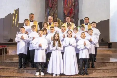 Dzieci w białych strojach komunijnych, z kapłanami, czekają na rozpoczęcie uroczystości. Zastanawiasz się, ile trwa msza?