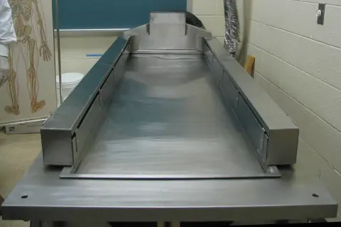 Stół do sekcji zwłok ze stali nierdzewnej, gotowy do pracy. Obok wisi tablica z anatomią człowieka.