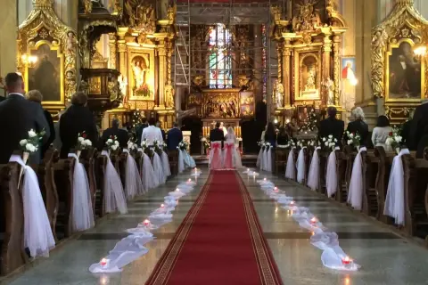 Wnętrze kościoła udekorowane na ślub. Wzdłuż czerwonego dywanu stoją ławki ozdobione białymi kwiatami i tiulem. Widać świece i gości.