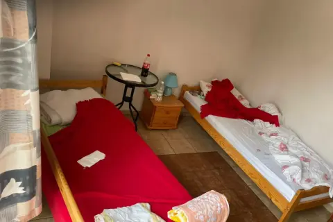 Pokój z dwoma łóżkami. Na jednym pościel po zmarłej osobie, przykryta czerwoną narzutą. Na drugim łóżku biała pościel z różowymi kwiatami i czerwony koc.