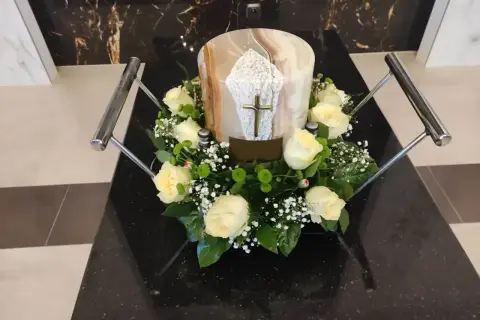 Białe róże i gipsówka otaczają urnę z krzyżem. Kwiaty na pogrzebie z urną symbolizują pamięć i szacunek.