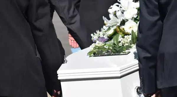 Pogrzeb członka rodziny, który nie był ubezpieczony. Biała trumna niesiona przez żałobników, ozdobiona bukietem białych kwiatów. Zasiłek pogrzebowy na nieubezpieczonego członka rodziny.