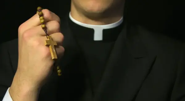 Dłoń księdza trzyma drewniany różaniec. Czy ksiądz może odmówić pochówku? Pytanie o sakramenty.