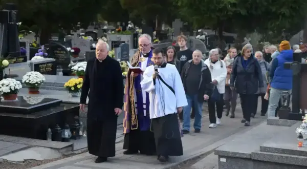 Ksiądz niesie urnę na pogrzebie, a za nim idzie grupa żałobników przez cmentarz.