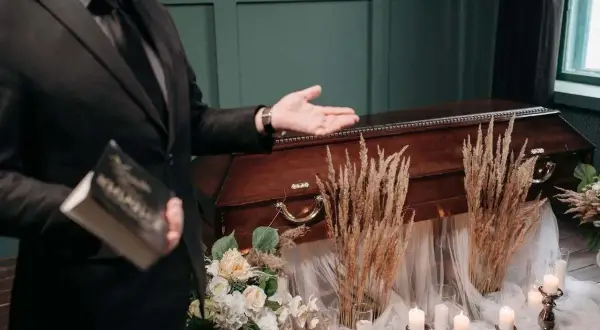 Mężczyzna w garniturze prezentuje trumnę, trzymając książkę. Scena sugeruje pogrzeby z zasadami, z kwiatami i świecami tworzącymi uroczystą atmosferę.