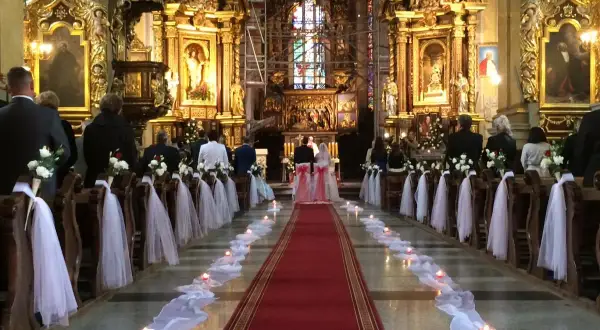 Wnętrze kościoła udekorowane na ślub. Wzdłuż czerwonego dywanu stoją ławki ozdobione białymi kwiatami i tiulem. Widać świece i gości.