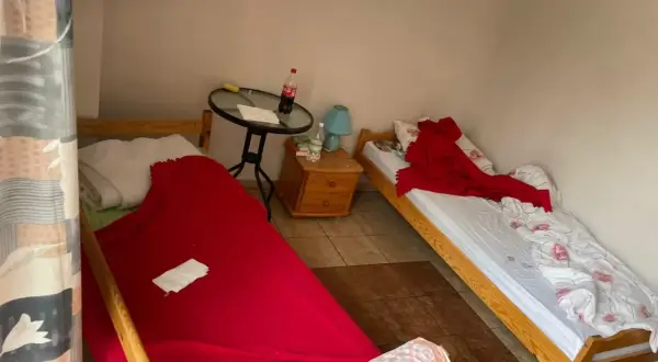 Pokój z dwoma łóżkami. Na jednym pościel po zmarłej osobie, przykryta czerwoną narzutą. Na drugim łóżku biała pościel z różowymi kwiatami i czerwony koc.