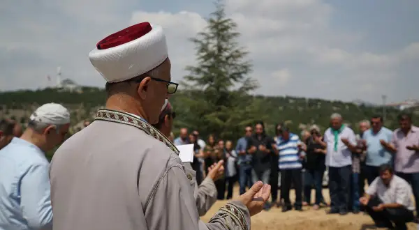 Uroczystość pogrzebowa muzułmańska. Duchowny w tradycyjnym stroju prowadzi modlitwę, zgromadzeni ludzie wznoszą ręce do nieba.