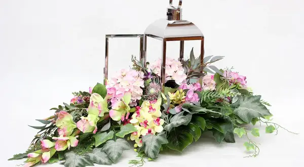 Elegancka kompozycja kwiatowa w lampionie, pełna różowych hortensji, żółtych i zielonych kwiatów, ozdobiona liśćmi i gałązkami.