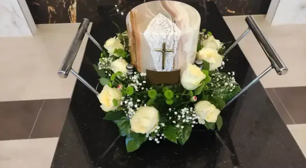 Białe róże i gipsówka otaczają urnę z krzyżem. Kwiaty na pogrzebie z urną symbolizują pamięć i szacunek.