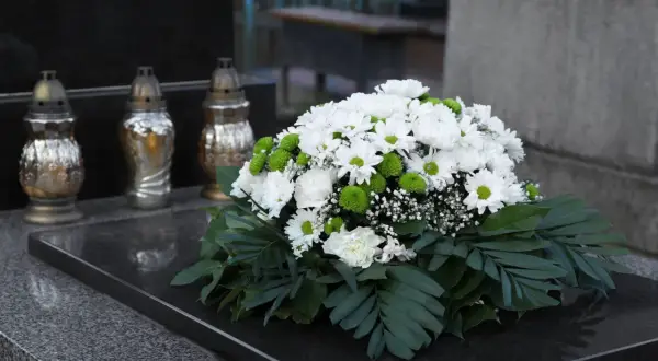 Białe i zielone kwiaty tworzą nowoczesne dekoracje na pomnik. Obok stoją trzy znicze.