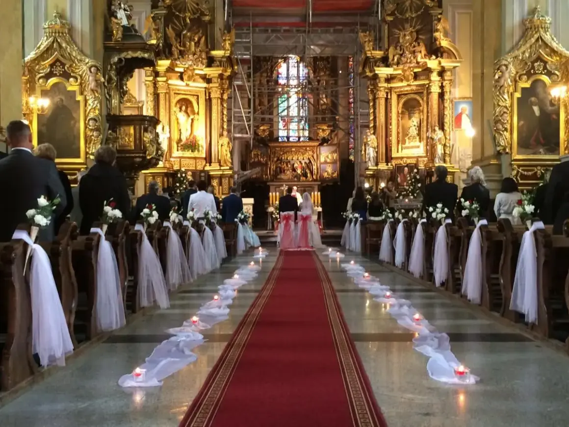 Wnętrze kościoła udekorowane na ślub. Wzdłuż czerwonego dywanu stoją ławki ozdobione białymi kwiatami i tiulem. Widać świece i gości.