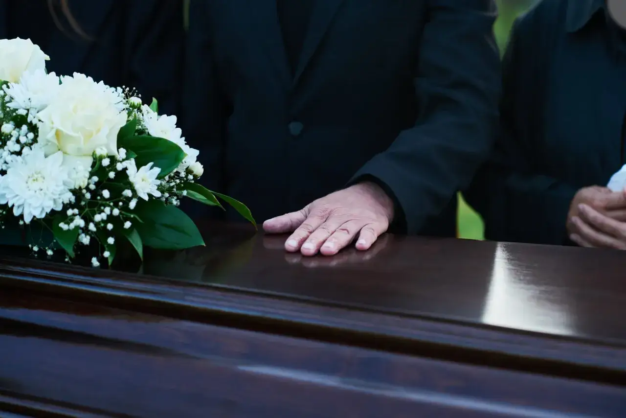 Dłoń spoczywa na ciemnej trumnie obok białych kwiatów. Uczestnicy w czerni podczas mszy pogrzebowej.