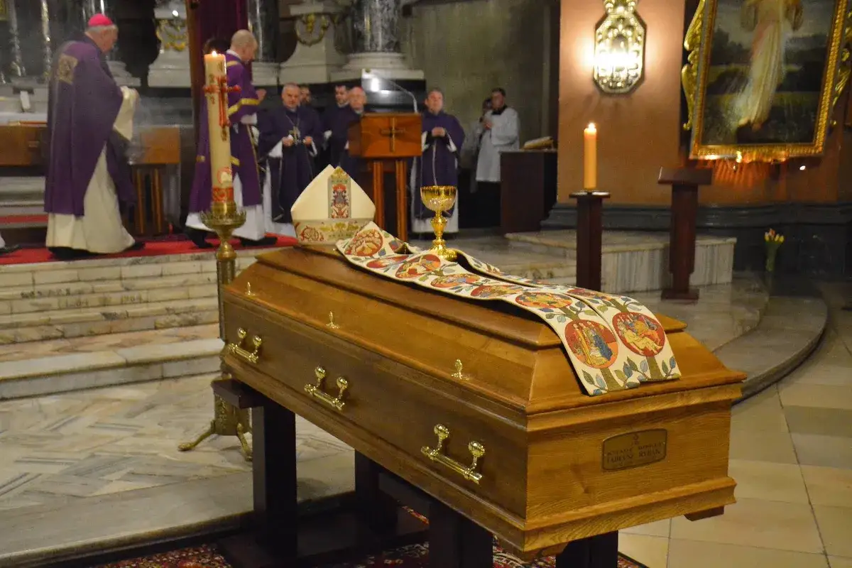 Drewniana trumna w kościele, ozdobiona stułą z biblijnymi scenami. Kapłani w strojach liturgicznych przygotowują się do mszy pogrzebowej krok po kroku.