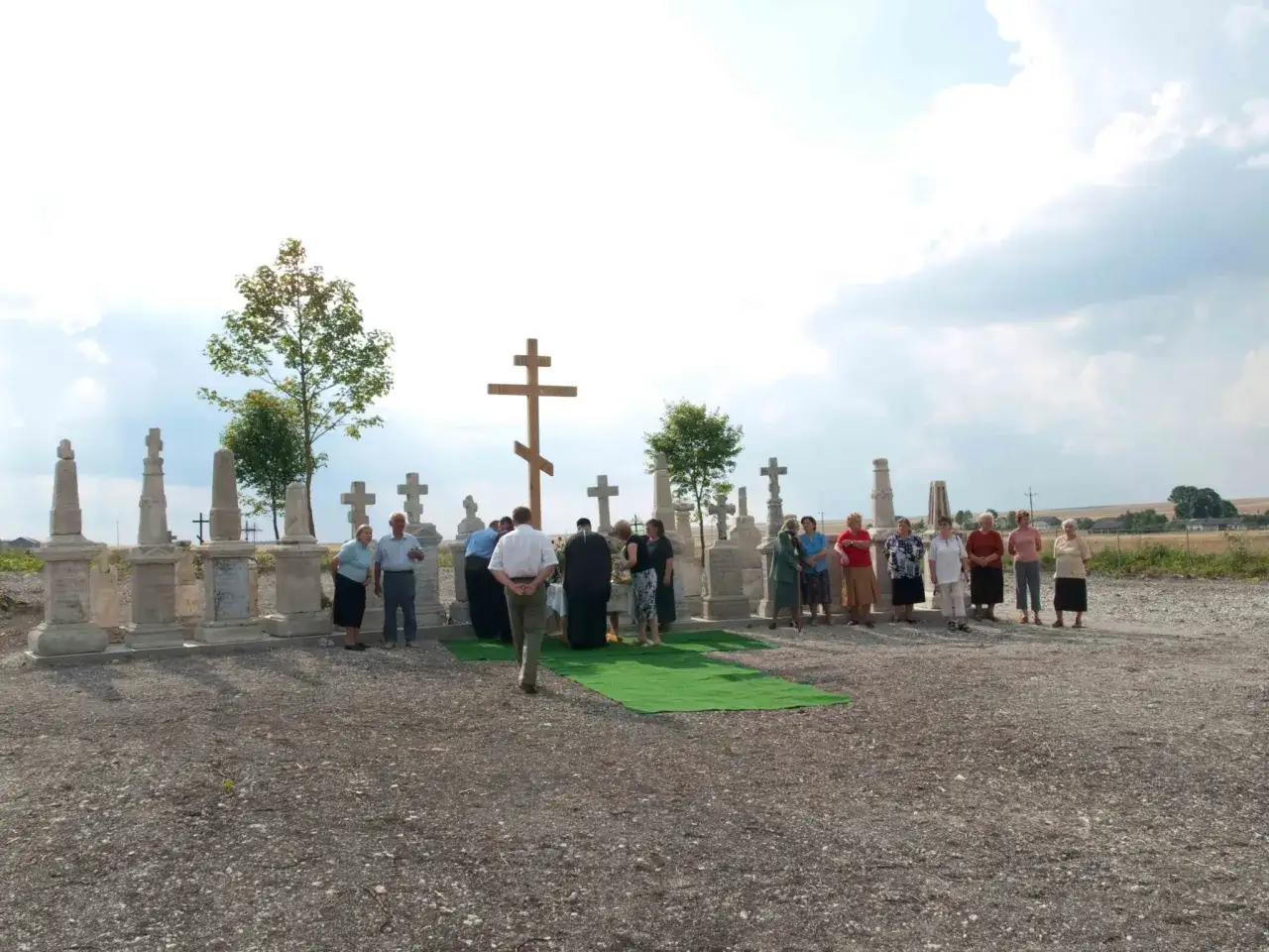 Uroczystość na cmentarzu, gdzie odmawia się modlitwę za zmarłych. Grupa ludzi zgromadzona wokół krzyża i stołu.