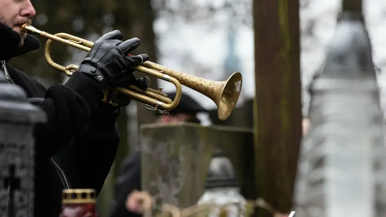 Trębacz w czarnej rękawiczce gra na cmentarzu. Dźwięk jego instrumentu wypełnia ciszę, tworząc przejmujący utwór pogrzebowy.