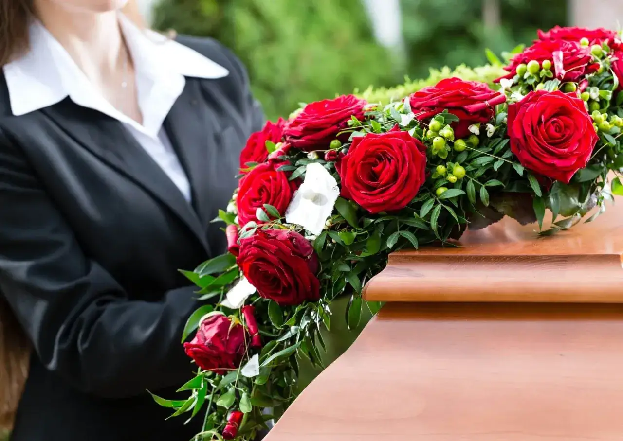 Osoba w czerni kładzie wieniec pogrzebowy dla mężczyzny, ozdobiony czerwonymi r&oacute;żami, na drewnianej trumnie.