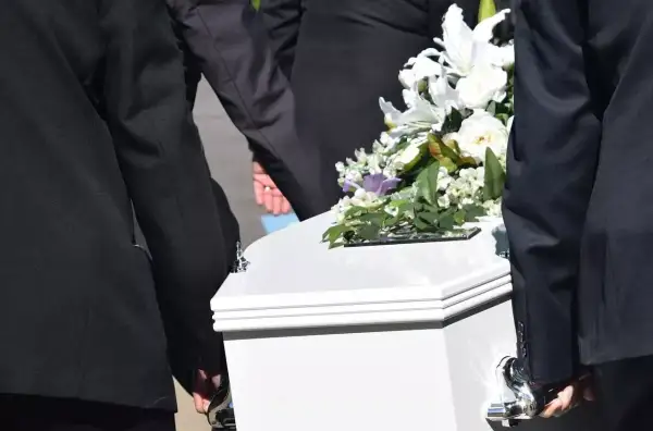 Pogrzeb członka rodziny, który nie był ubezpieczony. Biała trumna niesiona przez żałobników, ozdobiona bukietem białych kwiatów. Zasiłek pogrzebowy na nieubezpieczonego członka rodziny.