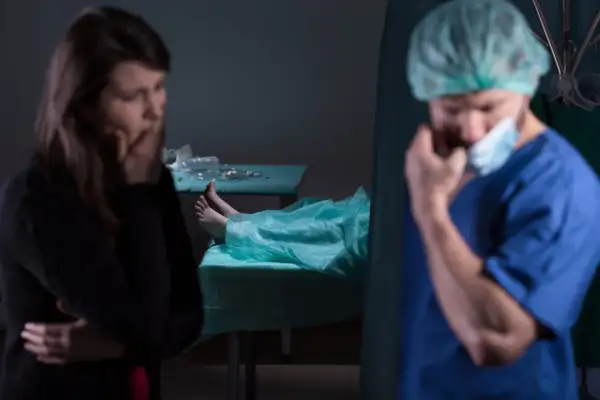Smutna kobieta i lekarz w masce, obok zakryte ciało na stole. Kiedy sekcja jest obowiązkowa, emocje biorą górę.